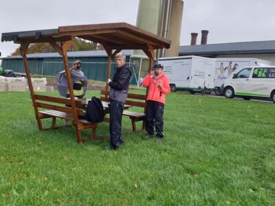 Drei Wanderteilnehmer stehen bei der Teepause unter einer Sitzgruppe mit Dach.