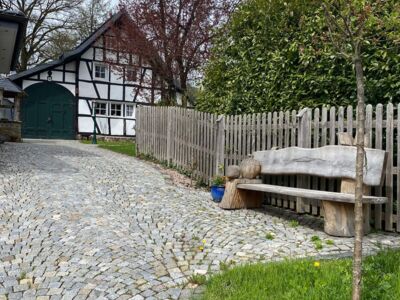 Das Foto zeigt eine Holzsitzbank an einem Zaun. Im Hintergrund ist ein Fachwerkhaus zu sehen.