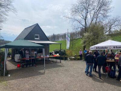 Auf dem Foto ist die SGV-Hütte mit Gartenzelten und die Verkaufsstation für Getränke, Kaffee und Kuchen zu sehen. Recht ist ein Stehtisch mit Besuchern.