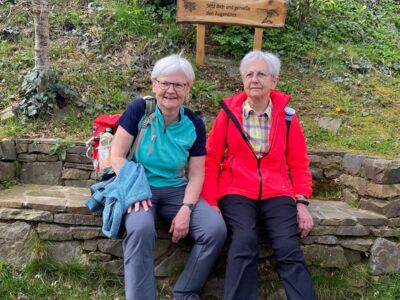 Auf dem Weg sind die beiden Wanderführerinnen zu sehen, die auf einer Steinbank Pause machen.